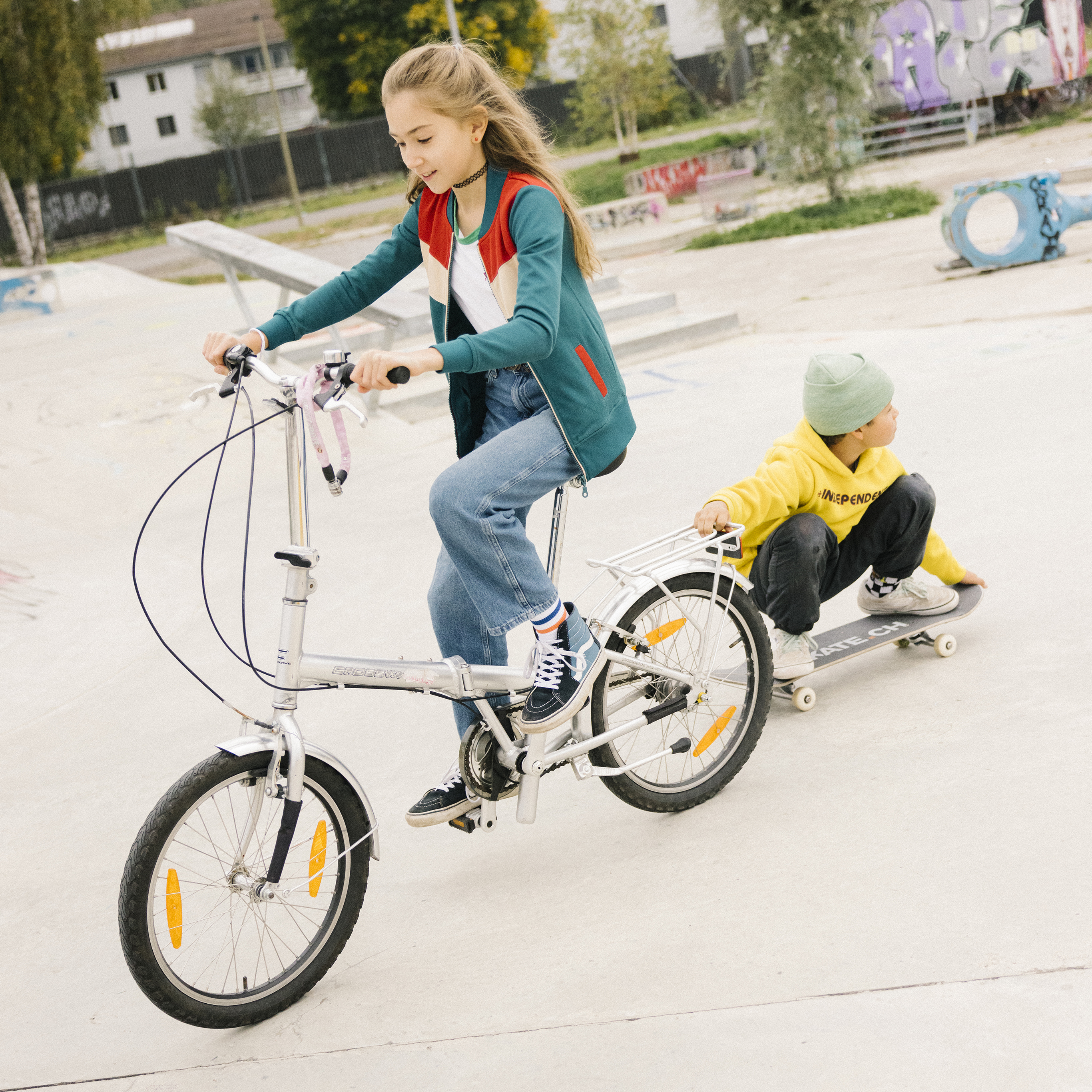 A teenager is riding her bicycle, while a teenager is crouching on his skateboard, holding onto the luggage rack and looking behind.