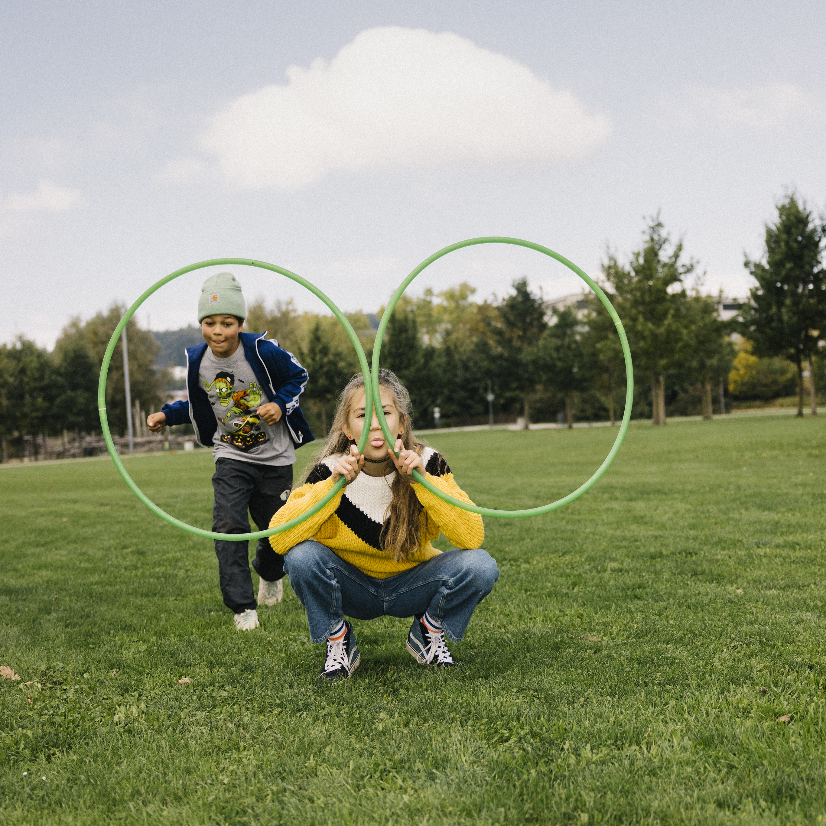 Eine Jugendliche hält zwei Hula-Hoop-Reifen wie eine Brille vor ihr Gesicht und streckt dabei die Zunge raus, während ein Jugendliche angerennt kommt, um durch einen der Ringe zu springen.