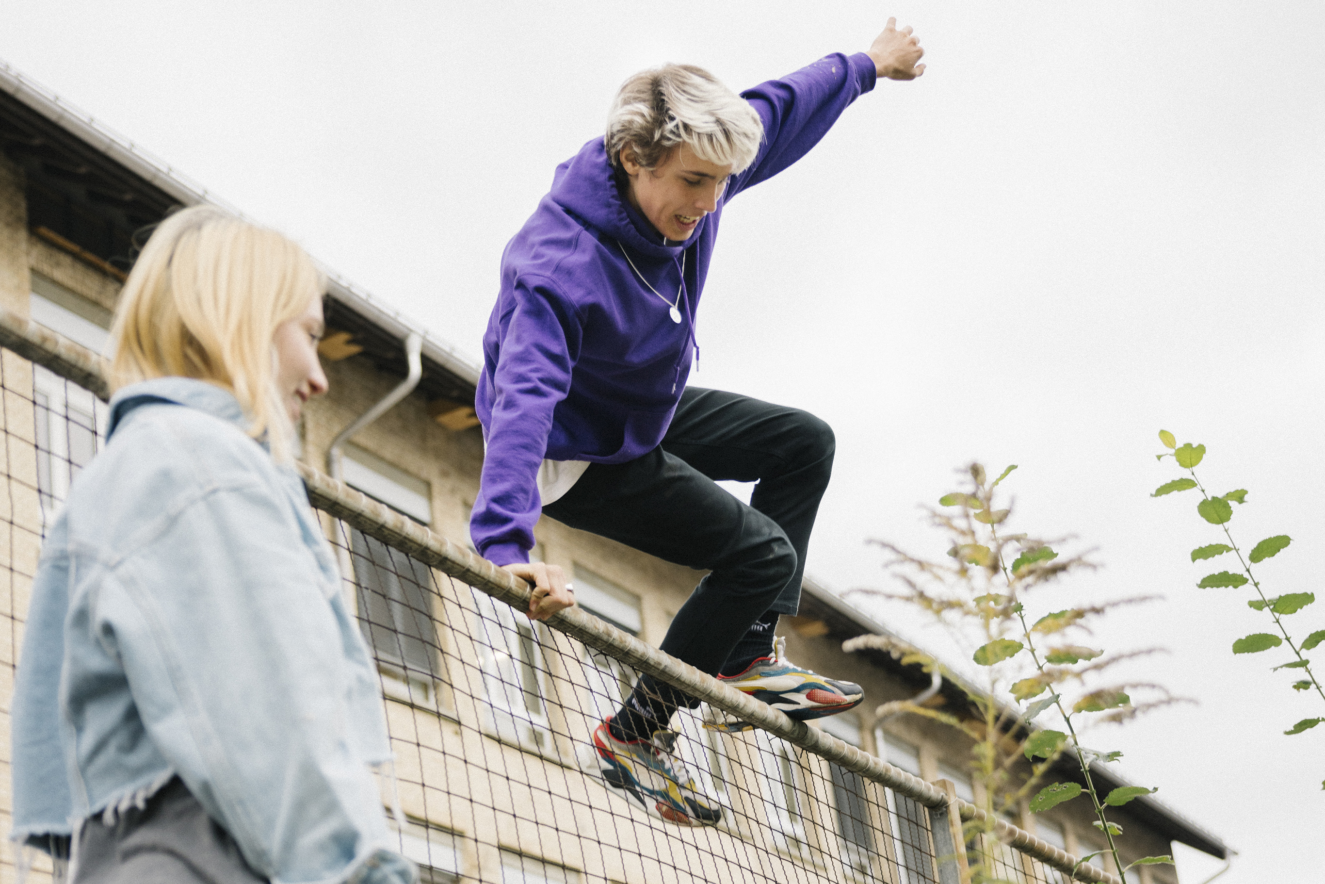 Young man jumps over high fence, observed by blond young woman. Both laugh. A building is visible in the background.
