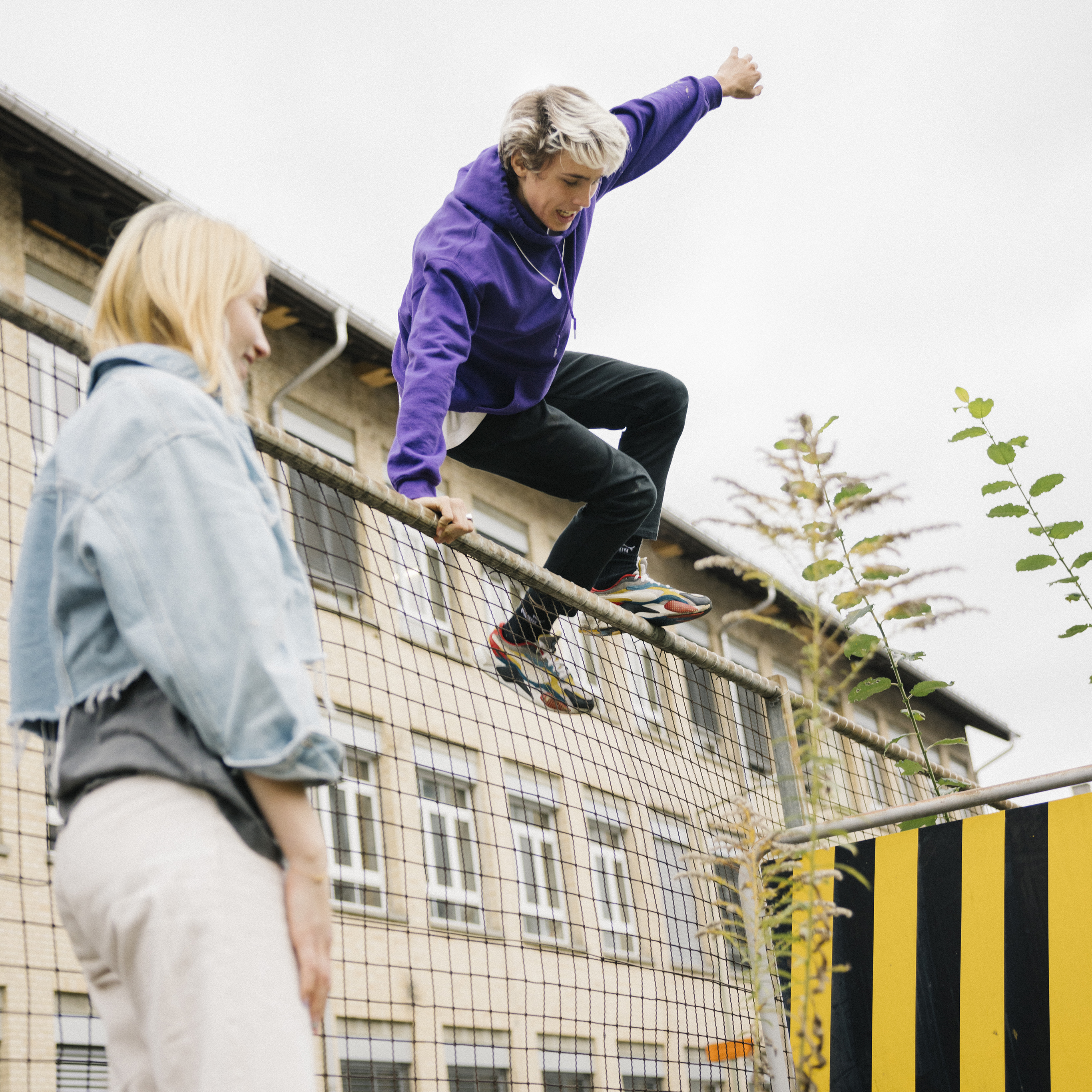 A young man jumps over a fence while a young woman looks happily but dreamily at the ground.