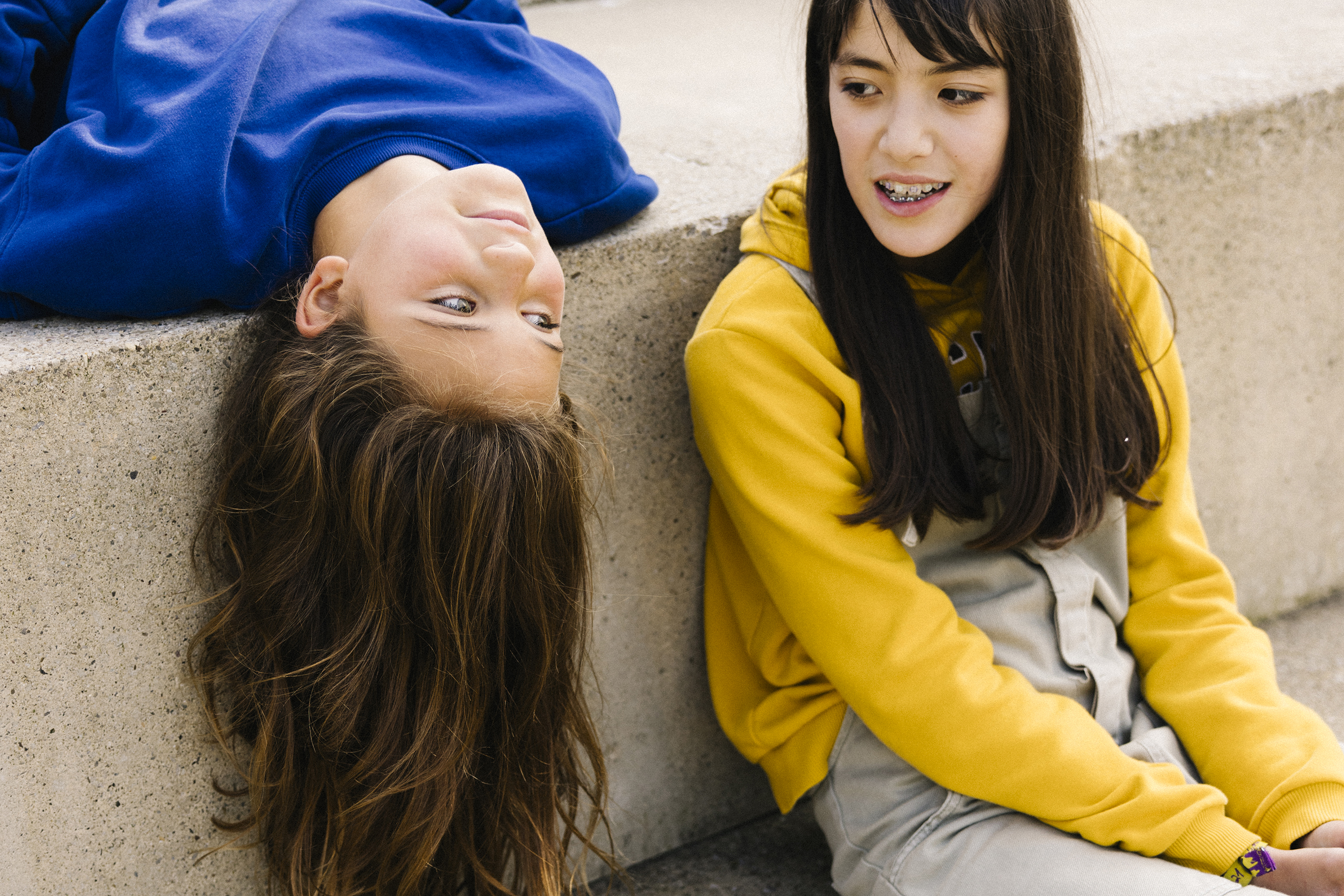 Two young women are talking on concrete steps, with one sittting on a step while another lies on the step on her back letting her head hang down.