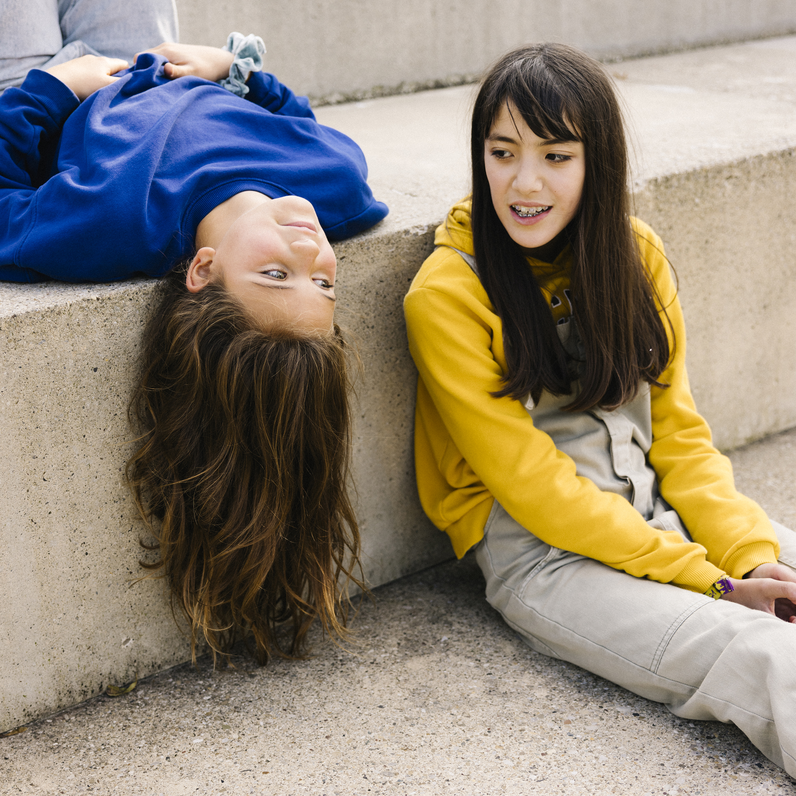 Deux adolescentes discutent sur un escalier en béton, l’une est assise sur une marche, tandis que l’autre allongée sur le dos laisse pendre sa tête sur le bord de la marche supérieure.