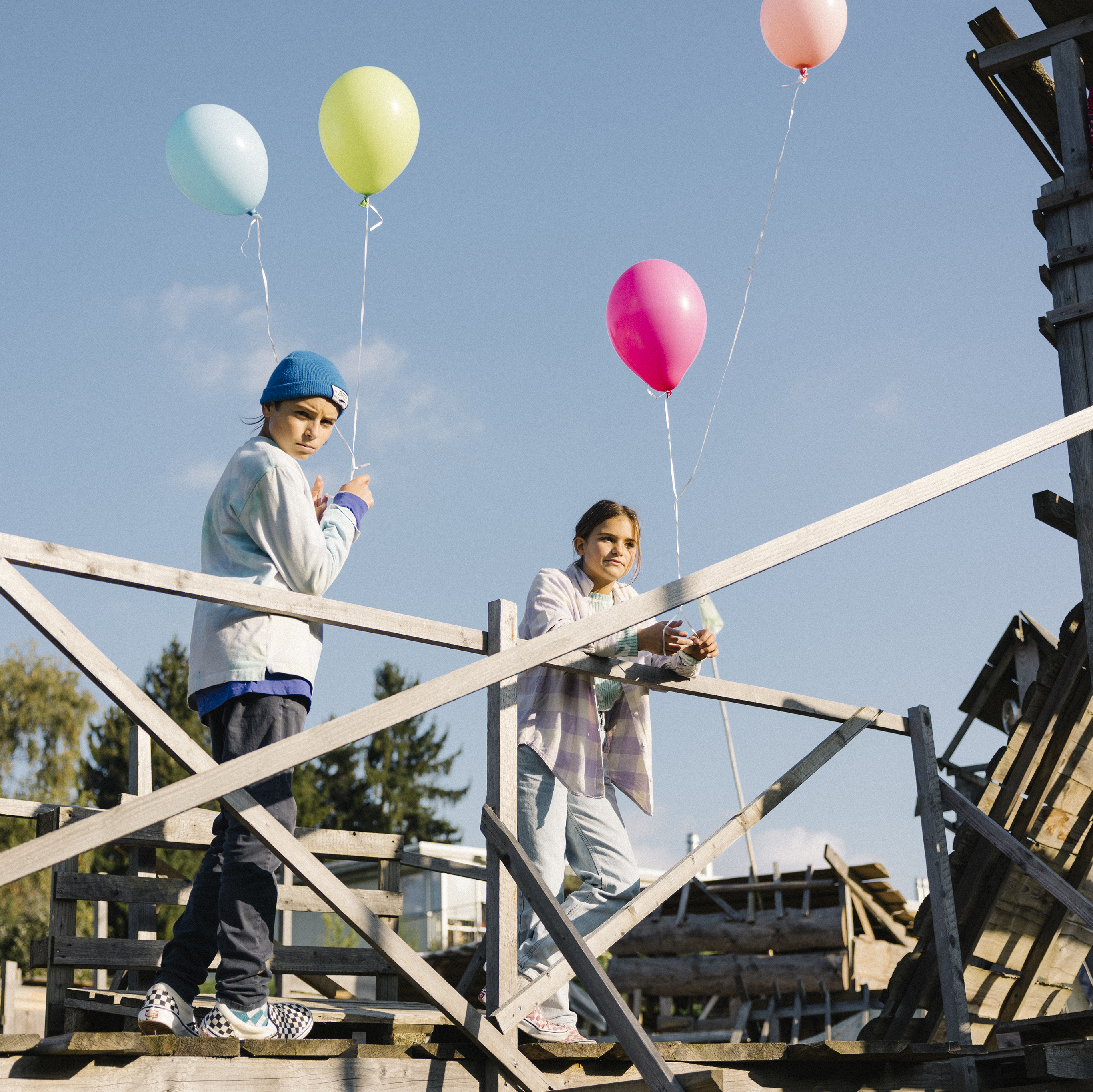 Deux adolescents appuyés contre une barrière le regard concentré tiennent chacun des fils reliés à deux ballons.