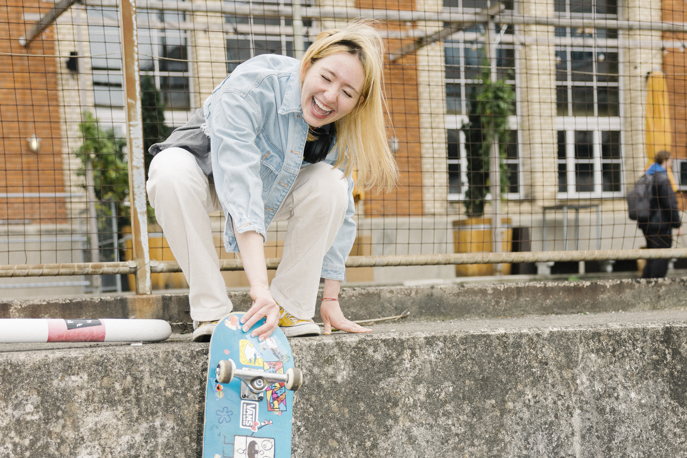 A young woman squats laughing on a wall holding her skateboard.