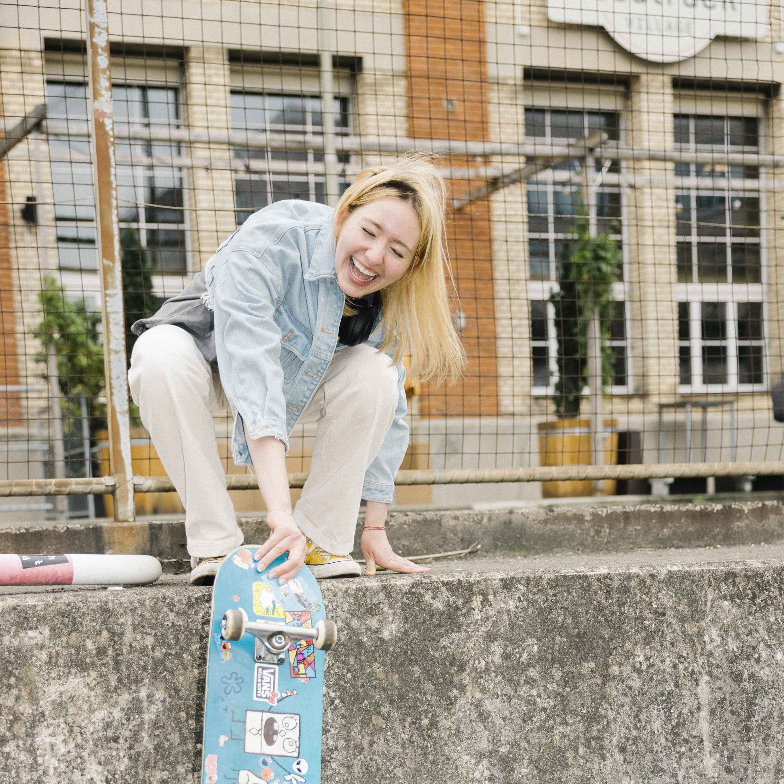 A teenager crouches on a wall laughing while holding her skateboard.