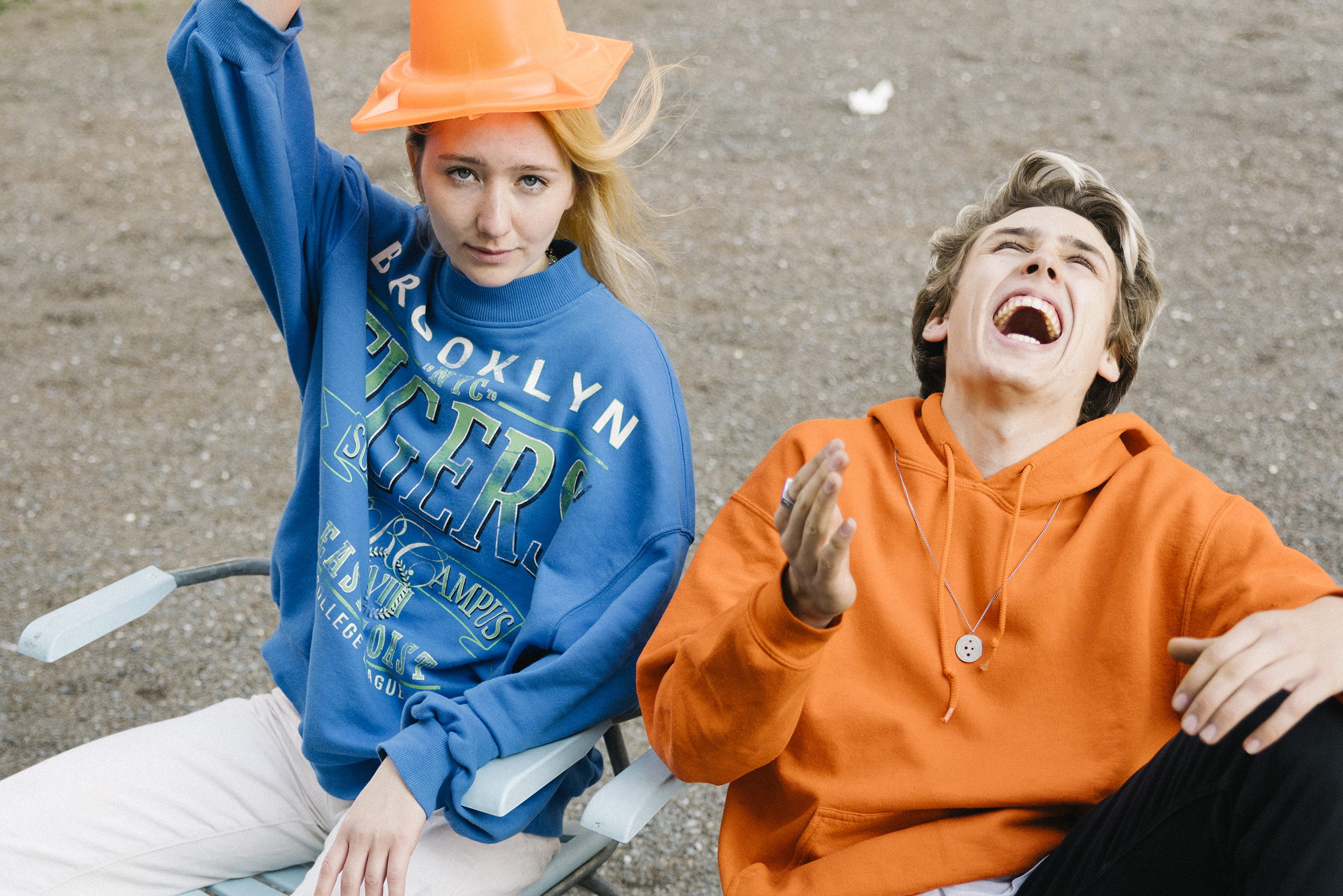 A youth laughs hysterically while looking up at the sky, while a youth looks into the camera while wearing a traffic cone as a hat.