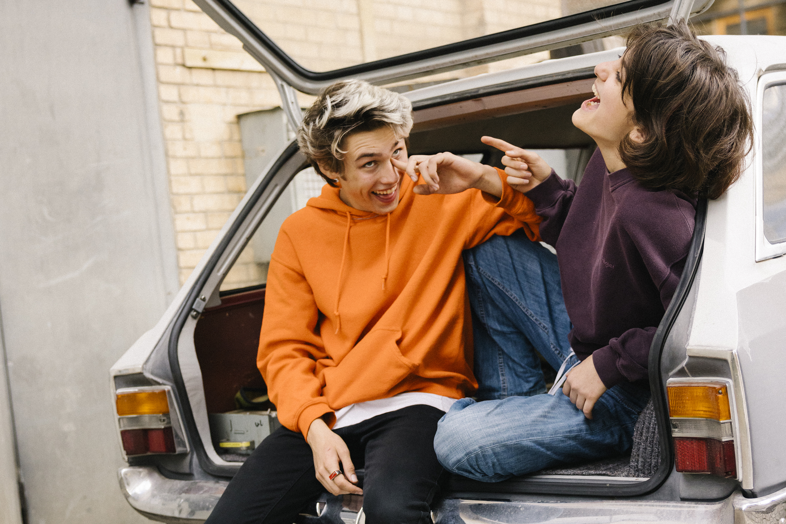 Two young people sit laughing in the open boot of a car.