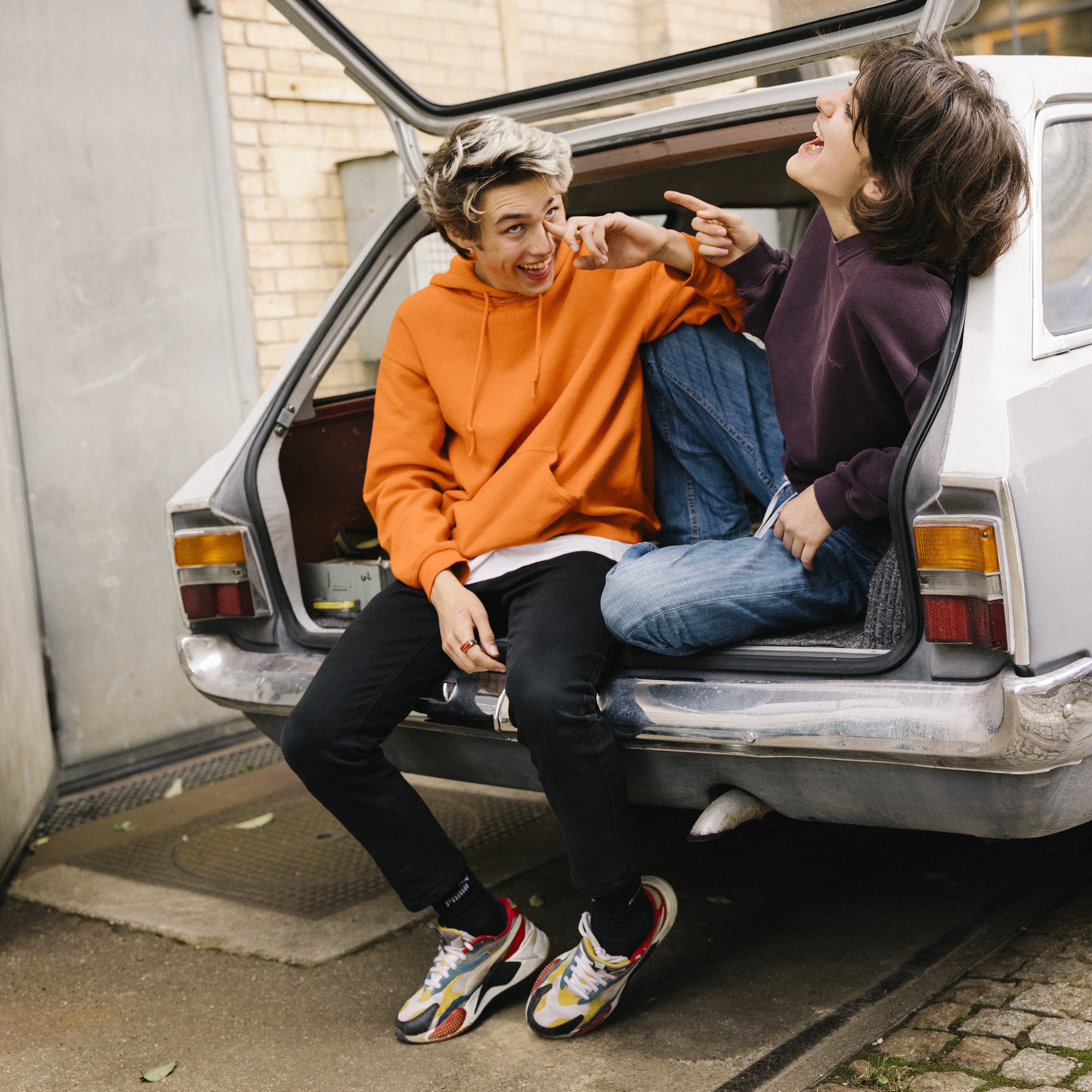 Two young people sit laughing in the open boot of a car.